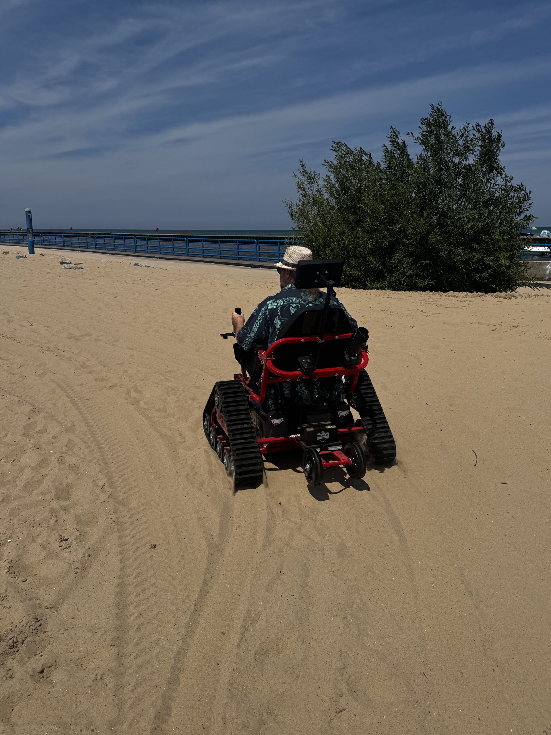 Bi-lateral above knee amputee driving a red and black powered chair with tank-treads on the beach.  The man is wearing a light colored fedora and a green and blue Hawaiian shirt, and is driving away, with his back to the camera.  