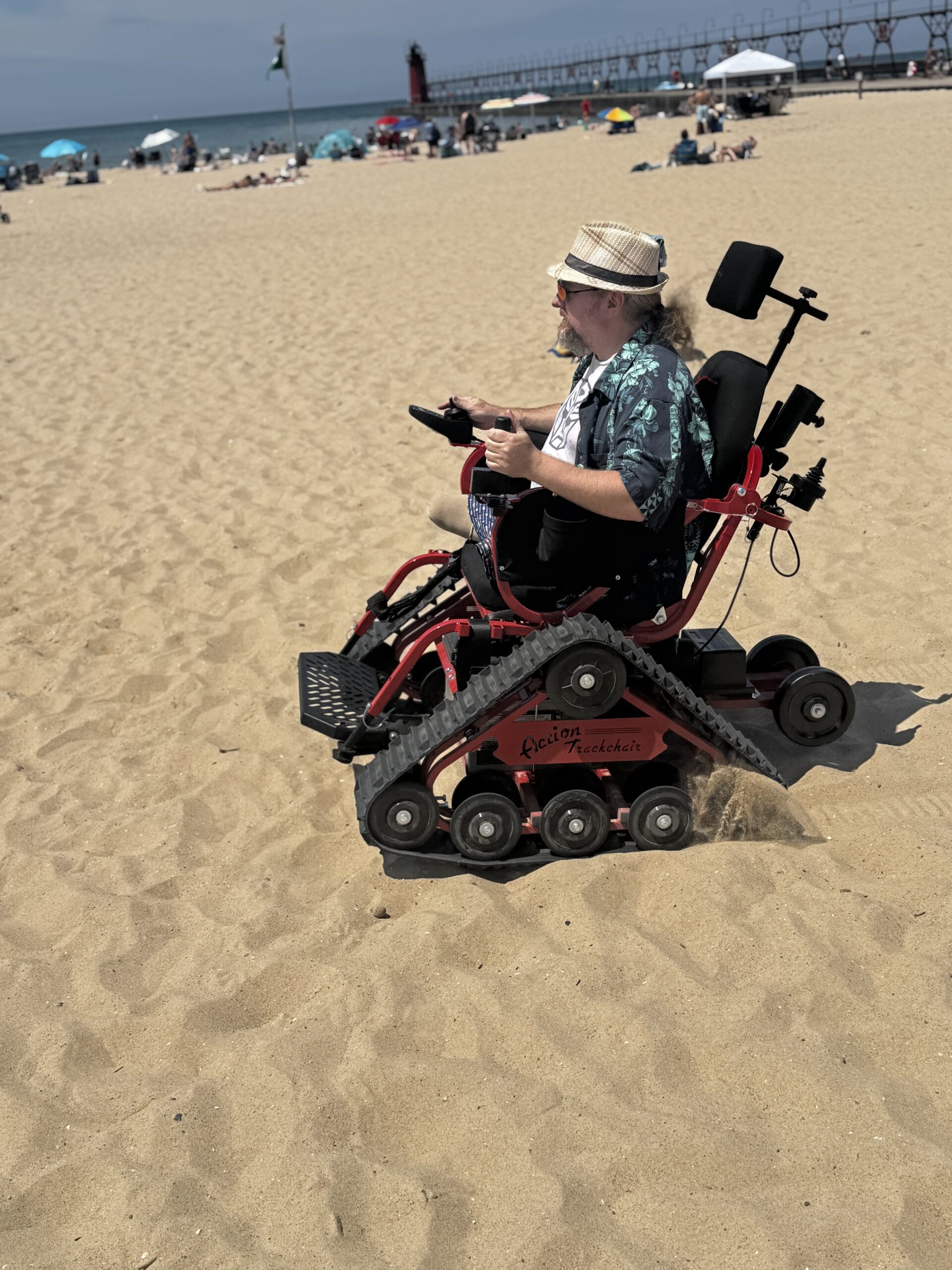 Bi-lateral above knee amputee driving a red and black powered chair with tank-treads on a sandy beach.  The man is wearing a light colored fedora, sunglasses, a t-shirt with an Autobot symbol in the middle, underneath an open button-down Hawaiian shirt, and a blue swimsuit. 