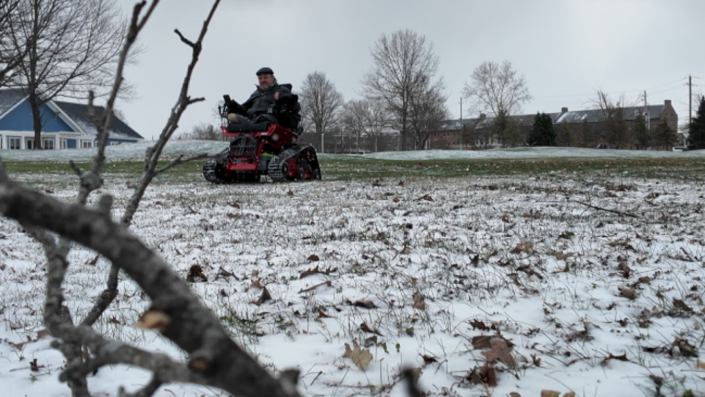 Bi-lateral above knee amputee driving a red and black chair with tank-treads on a grassy snow covered field.